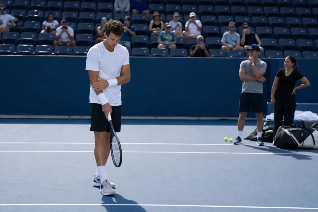Tennista in allenamento su campo in cemento blu mentre si tocca il polso, con spettatori e staff sullo sfondo