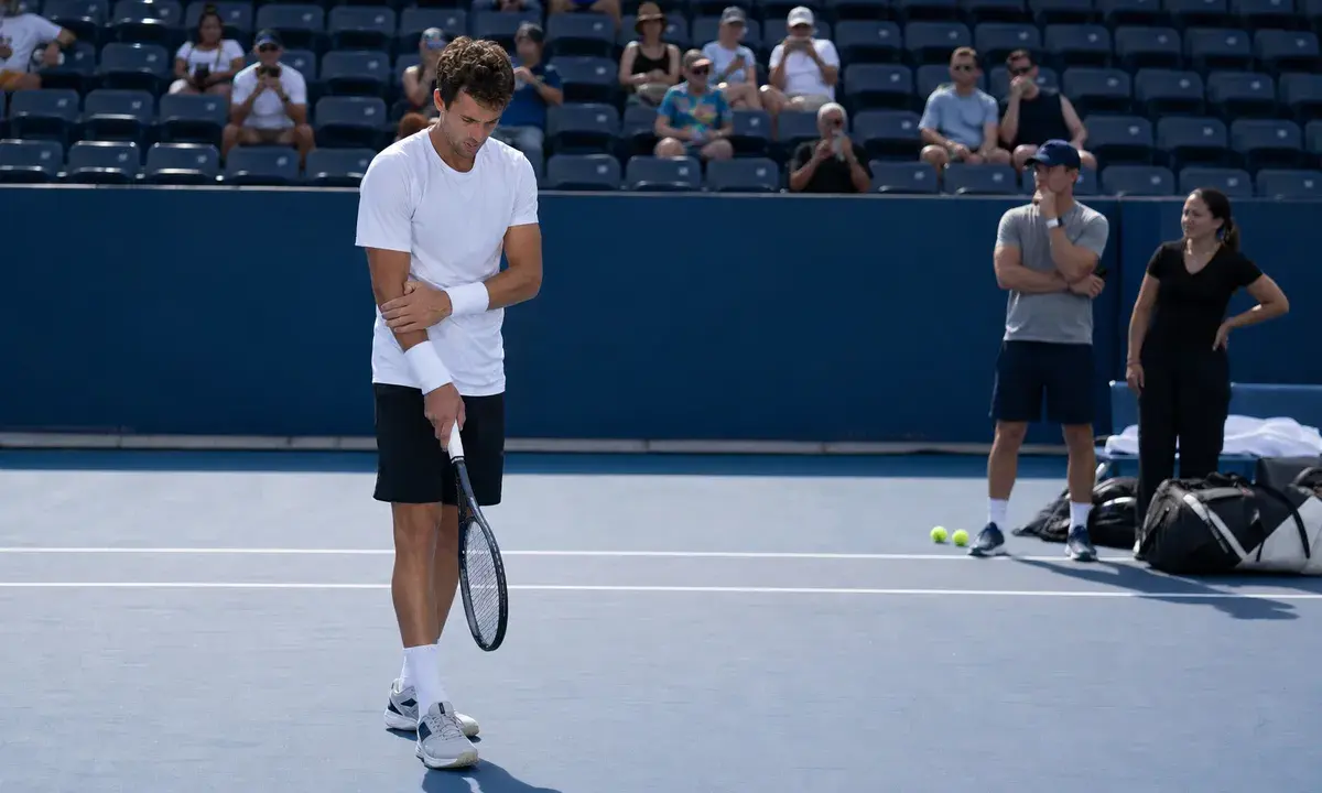 Tennista in allenamento su campo in cemento blu mentre si tocca il polso, con spettatori e staff sullo sfondo