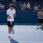 Tennista in allenamento su campo in cemento blu mentre si tocca il polso, con spettatori e staff sullo sfondo
