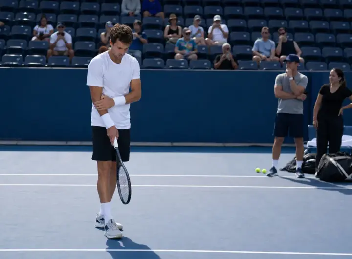 Tennista in allenamento su campo in cemento blu mentre si tocca il polso, con spettatori e staff sullo sfondo