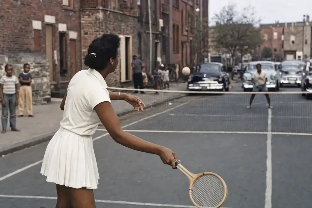 Donna in abito bianco gioca a tennis su un campo improvvisato in strada, con rete e auto d’epoca sullo sfondo