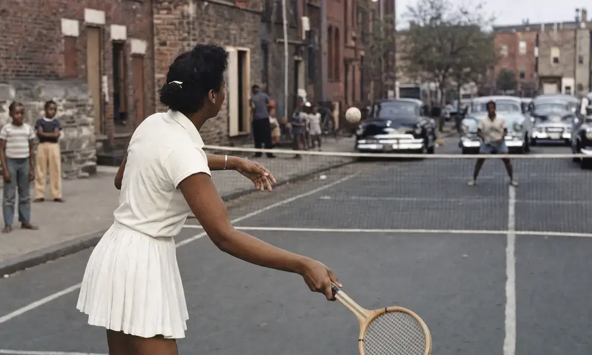 Donna in abito bianco gioca a tennis su un campo improvvisato in strada, con rete e auto d’epoca sullo sfondo