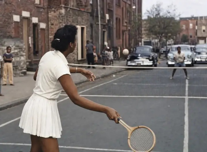 Donna in abito bianco gioca a tennis su un campo improvvisato in strada, con rete e auto d’epoca sullo sfondo