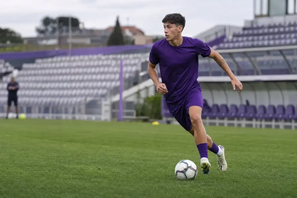 Giovane calciatore in completo viola durante l’allenamento, in corsa con il pallone su un campo da calcio