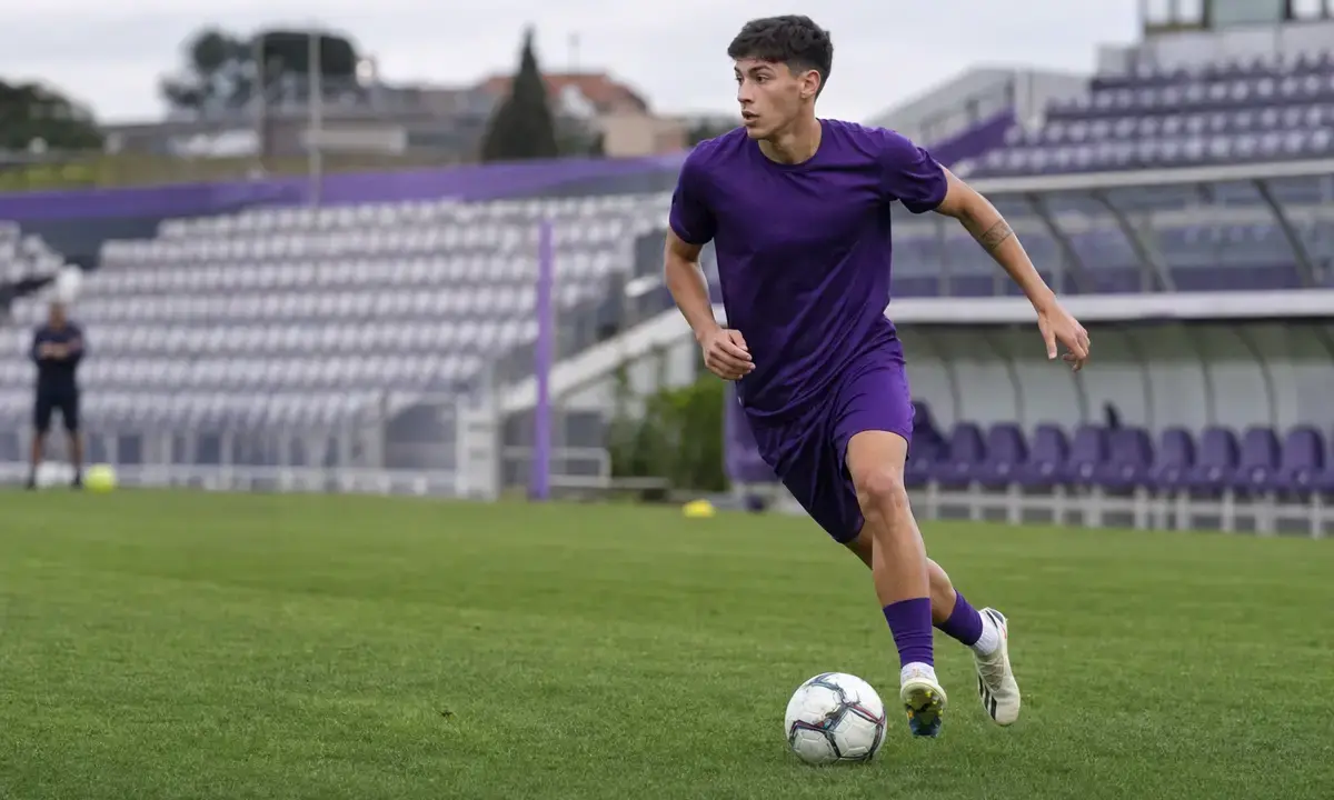 Giovane calciatore in completo viola durante l’allenamento, in corsa con il pallone su un campo da calcio