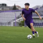 Giovane calciatore in completo viola durante l’allenamento, in corsa con il pallone su un campo da calcio