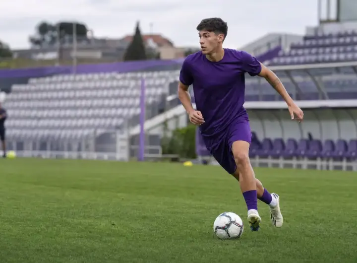 Giovane calciatore in completo viola durante l’allenamento, in corsa con il pallone su un campo da calcio