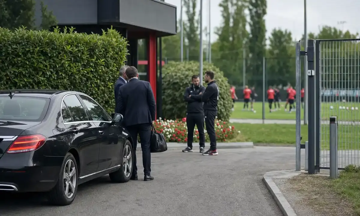 Auto nera all’ingresso di un centro sportivo, con dirigenti e staff che parlano e giocatori in rosso sul campo sullo sfondo