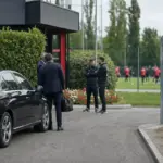 Auto nera all’ingresso di un centro sportivo, con dirigenti e staff che parlano e giocatori in rosso sul campo sullo sfondo