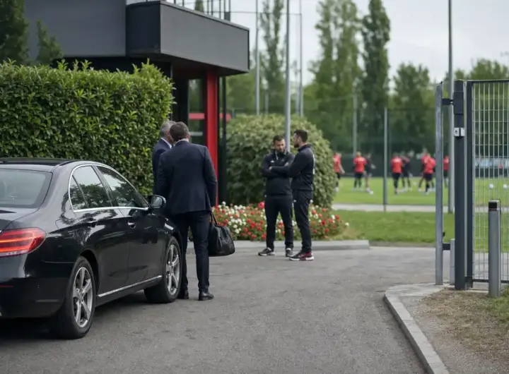 Auto nera all’ingresso di un centro sportivo, con dirigenti e staff che parlano e giocatori in rosso sul campo sullo sfondo