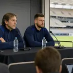 Allenatore e portiere in conferenza stampa allo stadio, seduti al tavolo con microfoni e bottiglie d’acqua