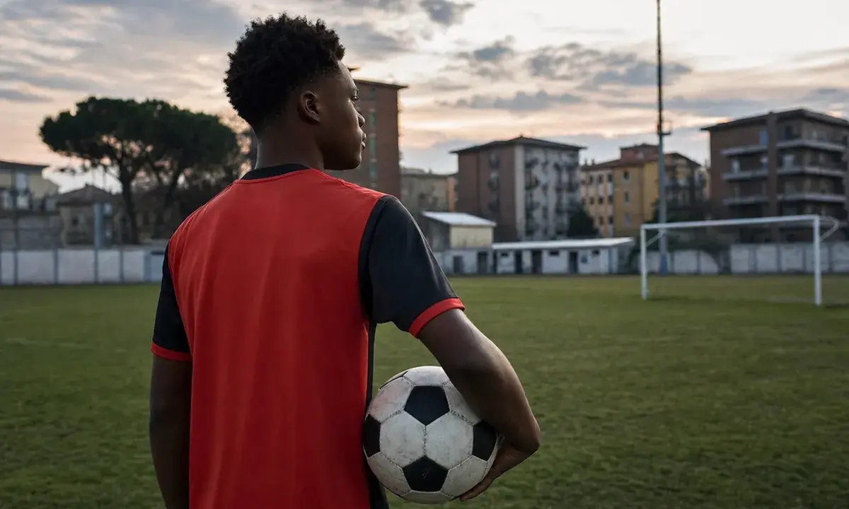 Giovane calciatore di spalle con maglia rossa e nera, pallone sotto braccio su un campo di periferia al tramonto