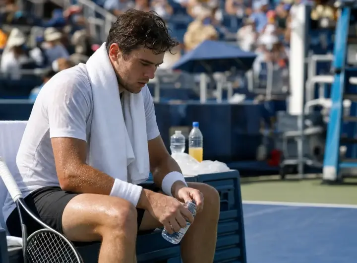 Tennista sudato e affaticato seduto in panchina con asciugamano al collo e bottiglietta d’acqua in mano