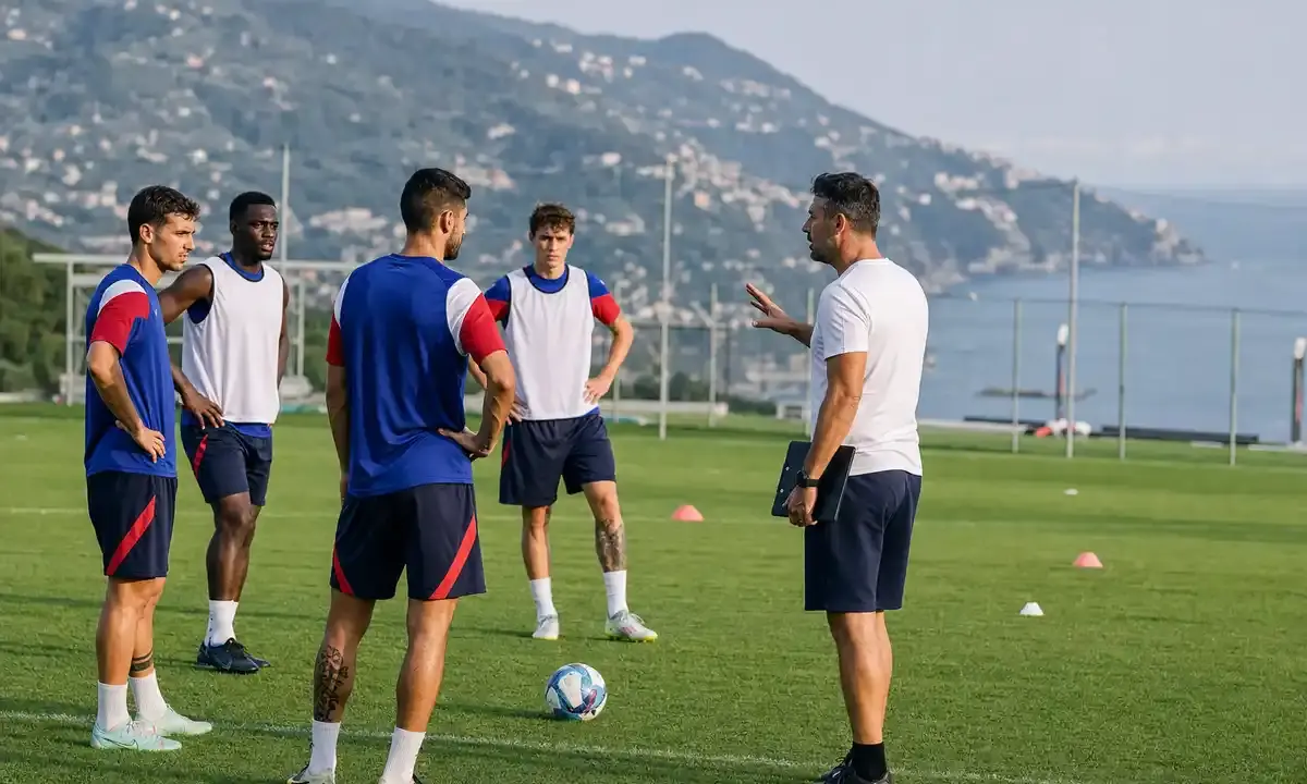 Allenatore parla a quattro calciatori in campo d’allenamento con pallone e coni, col mare sullo sfondo