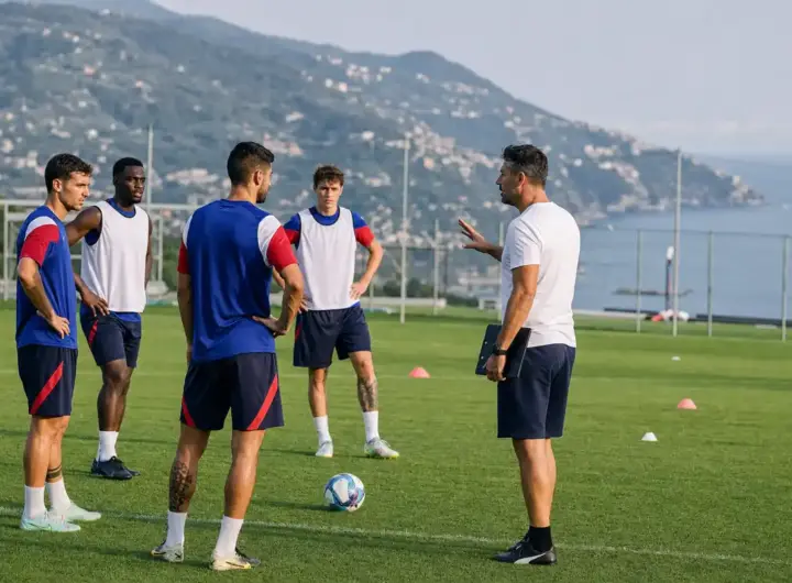 Allenatore parla a quattro calciatori in campo d’allenamento con pallone e coni, col mare sullo sfondo