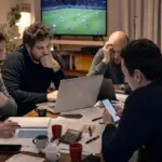 Quattro persone intorno a un tavolo con laptop, smartphone, fogli e calcolatrice durante un’asta di fantacalcio, TV sullo