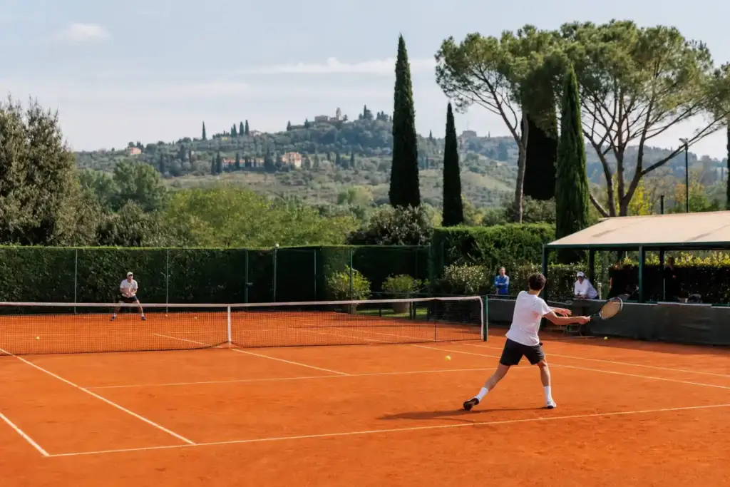 Giocatori in allenamento su campo da tennis in terra rossa con colline e cipressi sullo sfondo