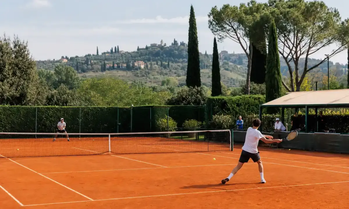 Giocatori in allenamento su campo da tennis in terra rossa con colline e cipressi sullo sfondo