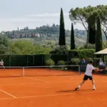 Giocatori in allenamento su campo da tennis in terra rossa con colline e cipressi sullo sfondo
