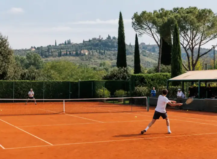 Giocatori in allenamento su campo da tennis in terra rossa con colline e cipressi sullo sfondo