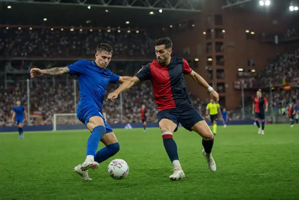 Contrasto a centrocampo tra due calciatori, uno in blu e uno in rosso e blu scuro, sotto i riflettori dello stadio