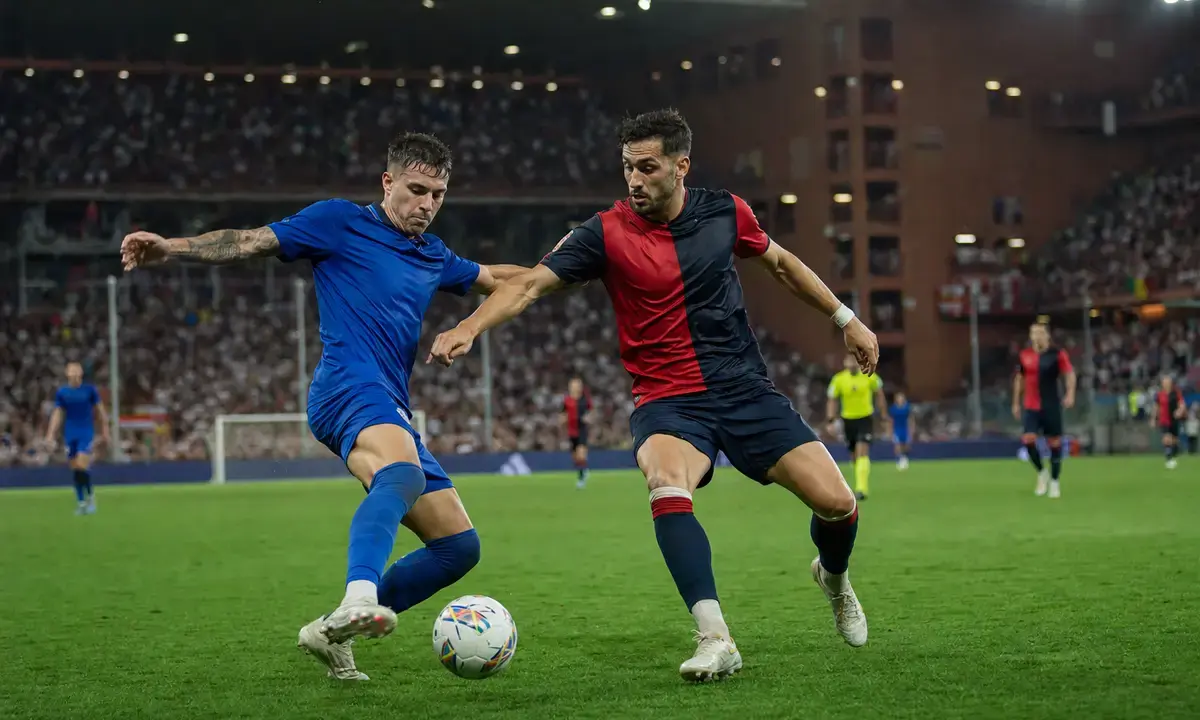 Contrasto a centrocampo tra due calciatori, uno in blu e uno in rosso e blu scuro, sotto i riflettori dello stadio