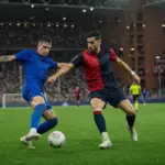 Contrasto a centrocampo tra due calciatori, uno in blu e uno in rosso e blu scuro, sotto i riflettori dello stadio