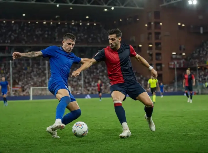 Contrasto a centrocampo tra due calciatori, uno in blu e uno in rosso e blu scuro, sotto i riflettori dello stadio