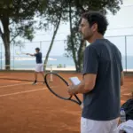 Allenatore con racchetta e taccuino osserva un giovane tennista in allenamento su campo in terra rossa sul mare