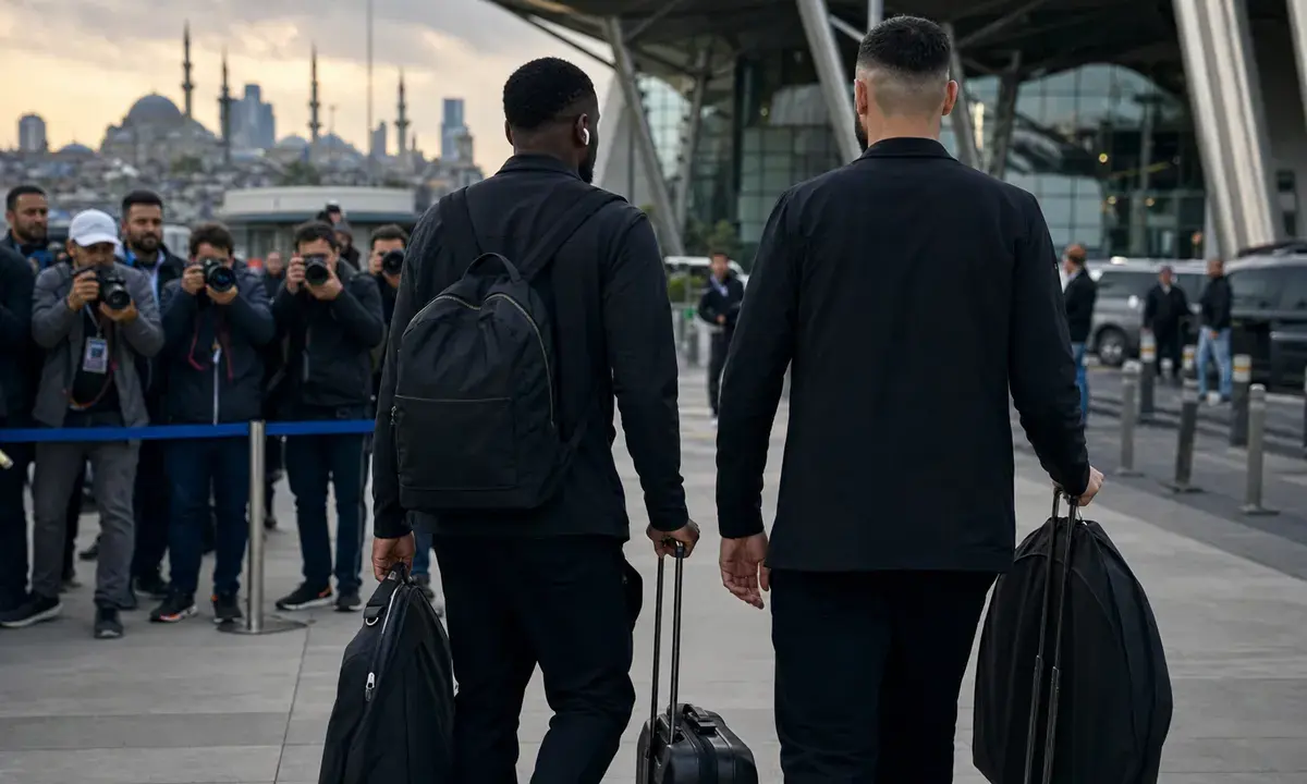 Due calciatori in abiti scuri, visti di spalle, trascinano trolley mentre fotografi li riprendono in un aeroporto