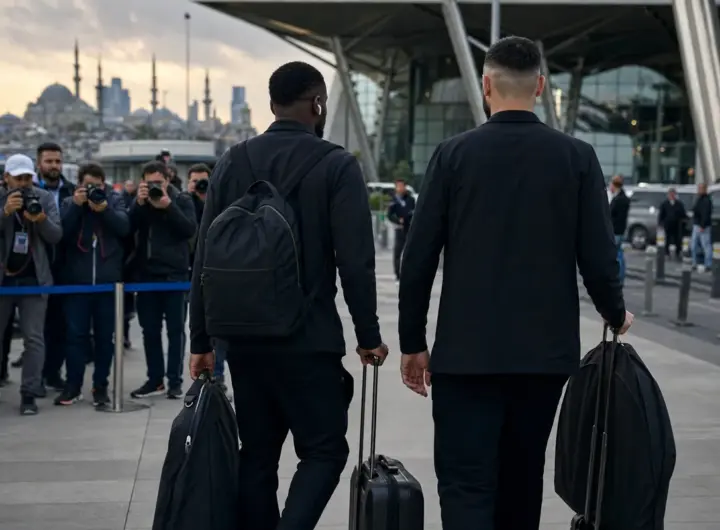 Due calciatori in abiti scuri, visti di spalle, trascinano trolley mentre fotografi li riprendono in un aeroporto
