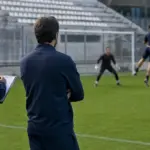 Allenatore e assistente a bordo campo osservano un portiere e due giocatori in allenamento con palla alta