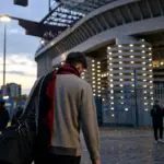 Calciatore di spalle con borsa e sciarpa rossonera che si allontana dallo stadio al tramonto