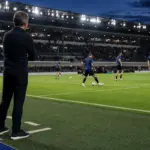 Allenatore a bordo campo osserva il riscaldamento di una squadra in maglia nerazzurra sotto i riflettori