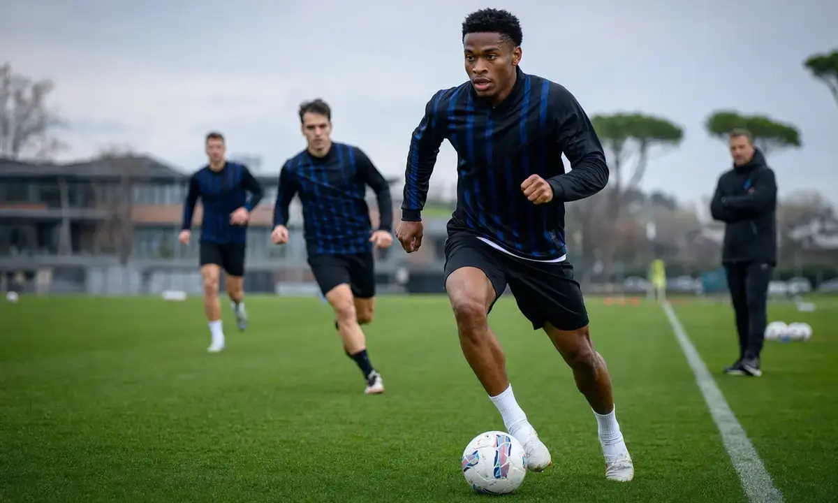 Calciatore in maglia scura a righe blu dribbla il pallone in allenamento, con compagni e allenatore sullo sfondo