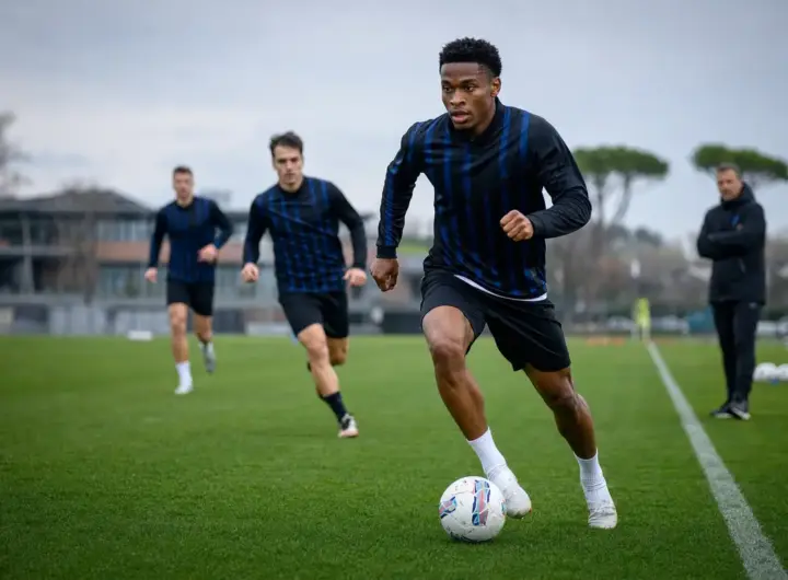 Calciatore in maglia scura a righe blu dribbla il pallone in allenamento, con compagni e allenatore sullo sfondo