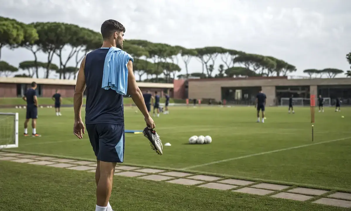 Calciatore di spalle entra in campo d’allenamento con scarpini in mano e maglia azzurra sulla spalla