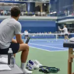 Tennista seduto a bordo campo con racchetta, mentre osserva un allenamento su un campo in cemento blu