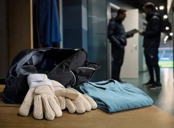 Guanti da portiere e borsa aperta su una panca, con maglia piegata; sullo sfondo due staff in corridoio