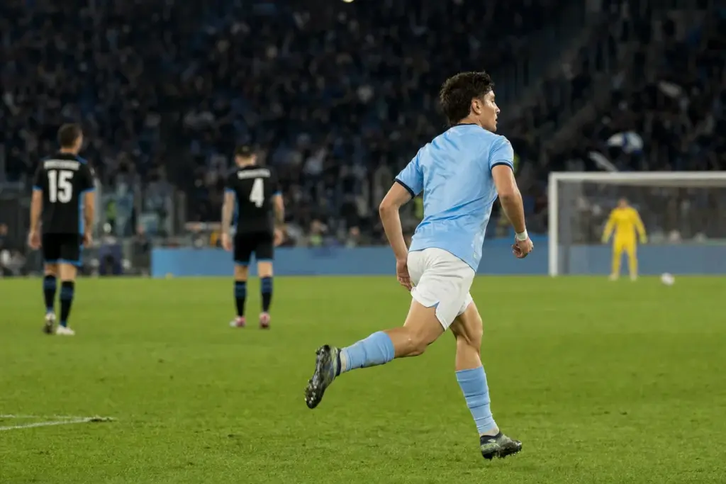 Centrocampista in maglia azzurro chiaro corre verso l’area di rigore in uno stadio illuminato di sera