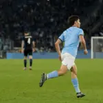 Centrocampista in maglia azzurro chiaro corre verso l’area di rigore in uno stadio illuminato di sera