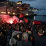 Tifosi con sciarpe e bandiere rosso-blu tra fumogeni, sullo sfondo il mare e le case di Boccadasse al tramonto