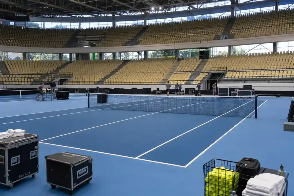 Campo da tennis indoor con superficie blu in un palazzetto vuoto, staff al lavoro e attrezzature a bordo campo
