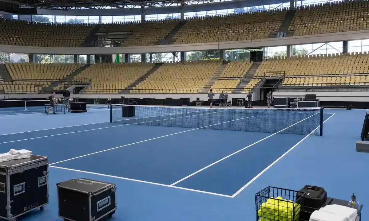 Campo da tennis indoor con superficie blu in un palazzetto vuoto, staff al lavoro e attrezzature a bordo campo
