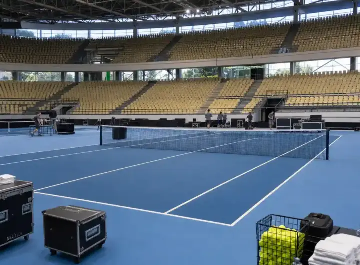 Campo da tennis indoor con superficie blu in un palazzetto vuoto, staff al lavoro e attrezzature a bordo campo