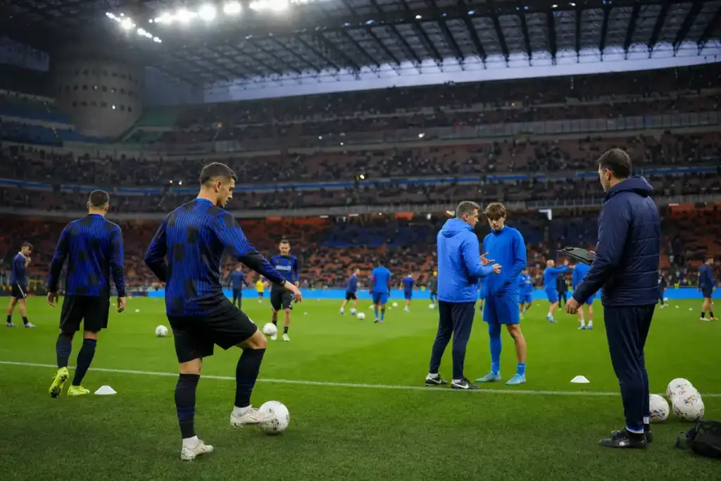 Riscaldamento prepartita in uno stadio illuminato, con giocatori in maglie scure a righe e in tuta blu, palloni e coni