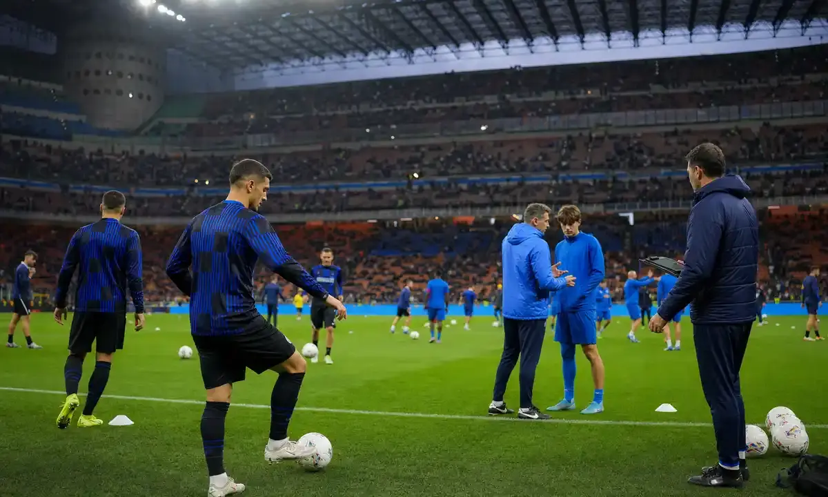 Riscaldamento prepartita in uno stadio illuminato, con giocatori in maglie scure a righe e in tuta blu, palloni e coni