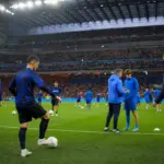Riscaldamento prepartita in uno stadio illuminato, con giocatori in maglie scure a righe e in tuta blu, palloni e coni