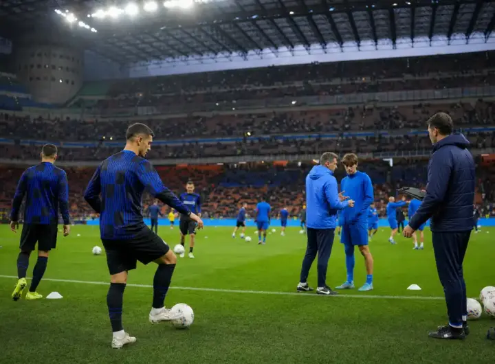 Riscaldamento prepartita in uno stadio illuminato, con giocatori in maglie scure a righe e in tuta blu, palloni e coni