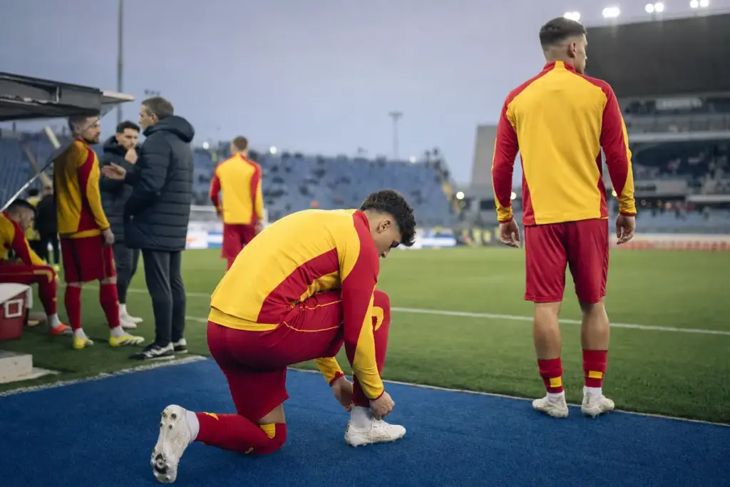 Calciatori in tuta giallorossa a bordo campo: uno si allaccia gli scarpini, un altro guarda il terreno di gioco